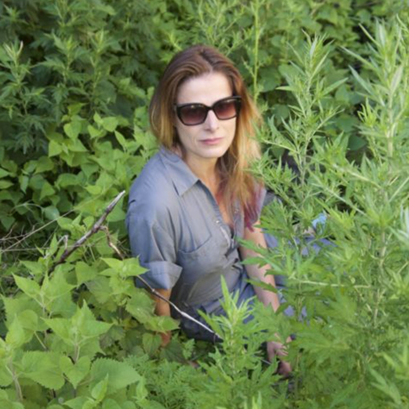 A photo f andrea wearing a blue long sleeve collared shirt and sunglasses immersed in greenery.